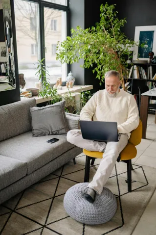 Elderly man using a laptop indoors with natural light and indoor plants.
