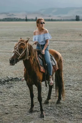 A woman in a bardot top rides a horse through an open field, enjoying the countryside view.