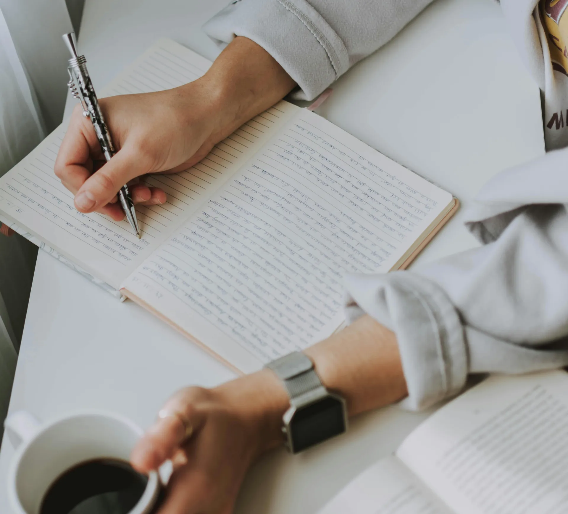 Close-up of a person writing in a notebook with coffee on a desk. Ideal for workplace or study themes.