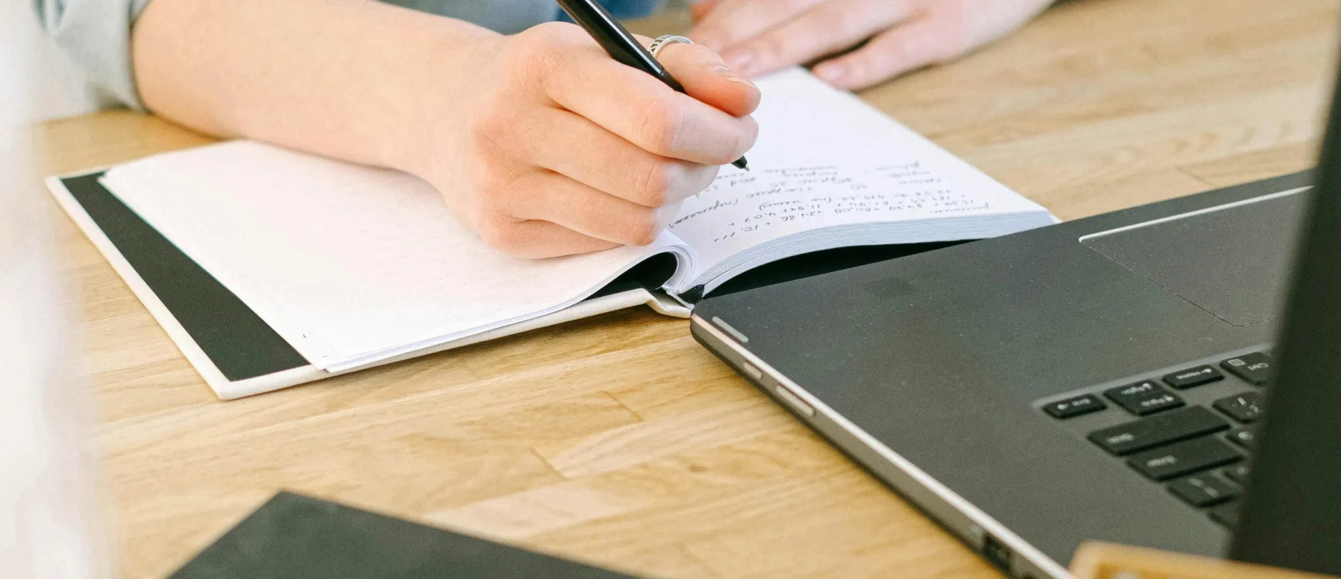 Adult writing notes on a notebook while working on a laptop in a cozy home office environment.