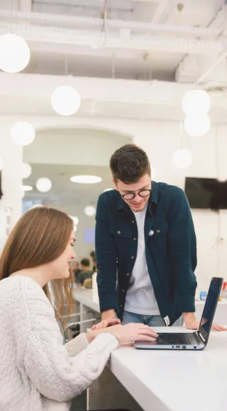 Two young adults collaborating over a laptop in a modern, bright office setting.