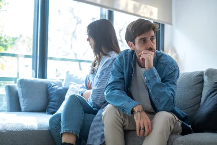 Young millennial couple sitting back to back on a sofa at home, sad and upset, ignoring each other, struggling with rela