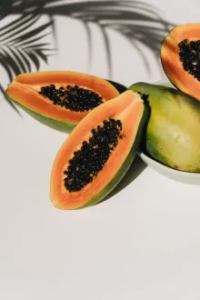 From above of delicious papaya halves with whole fruit placed on white surface in white round plate under shadow of palm leaves