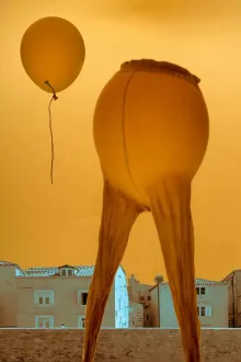 A whimsical scene featuring balloons against an orange sunset sky over a cityscape, creating a surreal atmosphere.