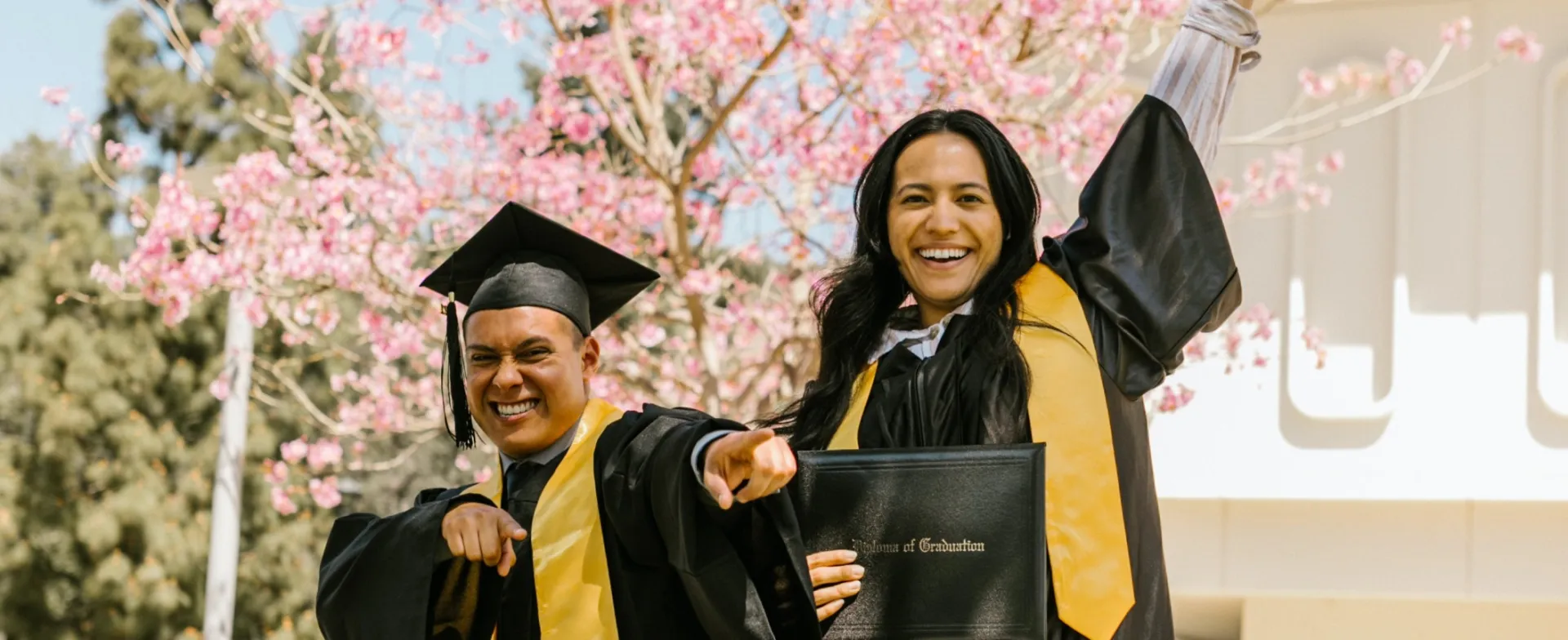 A Man and Woman in Black Academic Regalia Smiling while Holding a Diploma