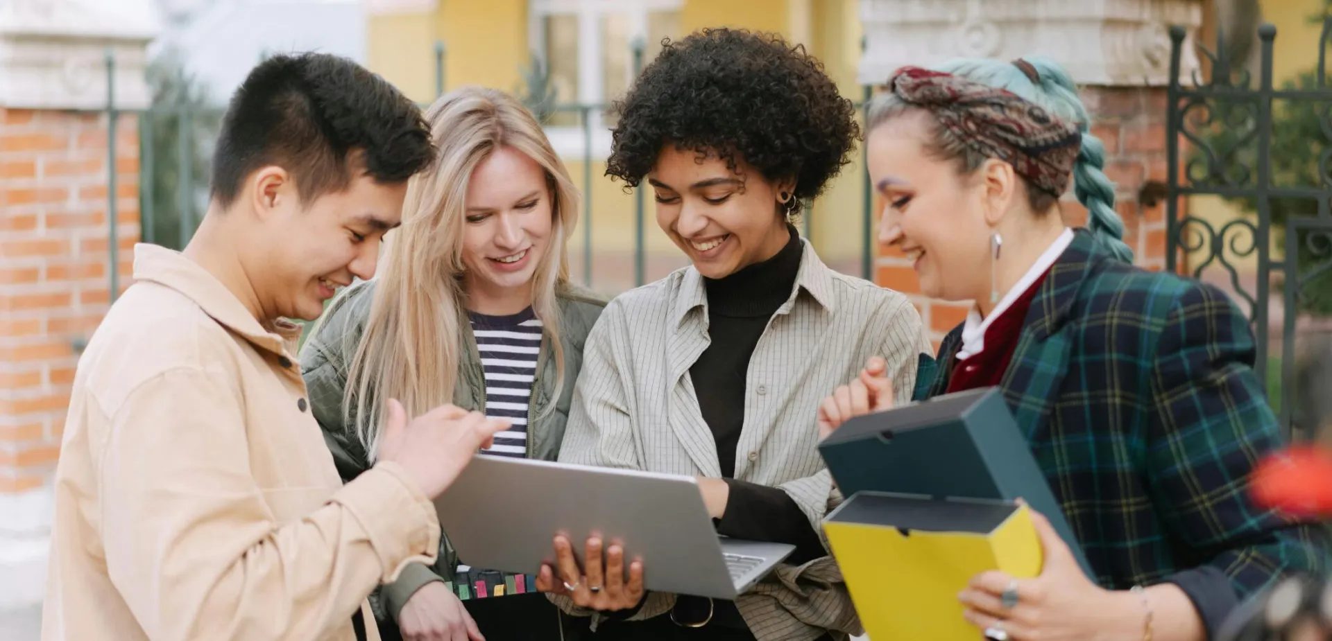 A diverse group of young adults smiling and collaborating outdoors with a laptop.