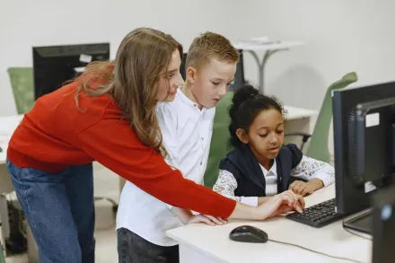 Young teacher helps students with a computer task in a classroom setting.
