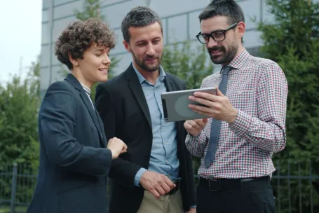 Three professionals engaged in a discussion while viewing a tablet outdoors.