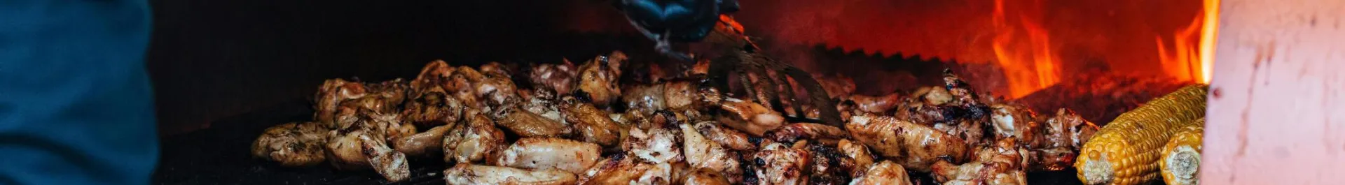 Close-up of grilling chicken wings and corn on an outdoor barbecue with flames.