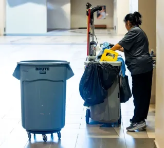 A janitorial worker pushes a cleaning cart in a spacious indoor hallway.