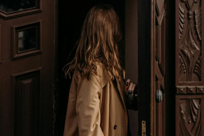 Woman in a brown coat entering through a classic ornate wooden door, creating a fashion narrative.