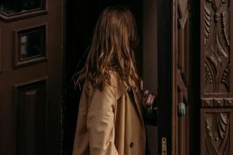 Woman in a brown coat entering through a classic ornate wooden door, creating a fashion narrative.