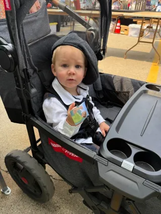 Young boy dressed smartly including a cap in stroller holding a sticker from Into the Thicket, picture book.