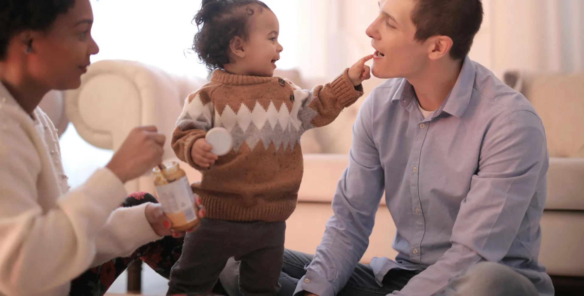 Side view of diverse parents in casual clothing sitting on floor and having fun with smiling toddler while gathering in cozy living room and having rest together