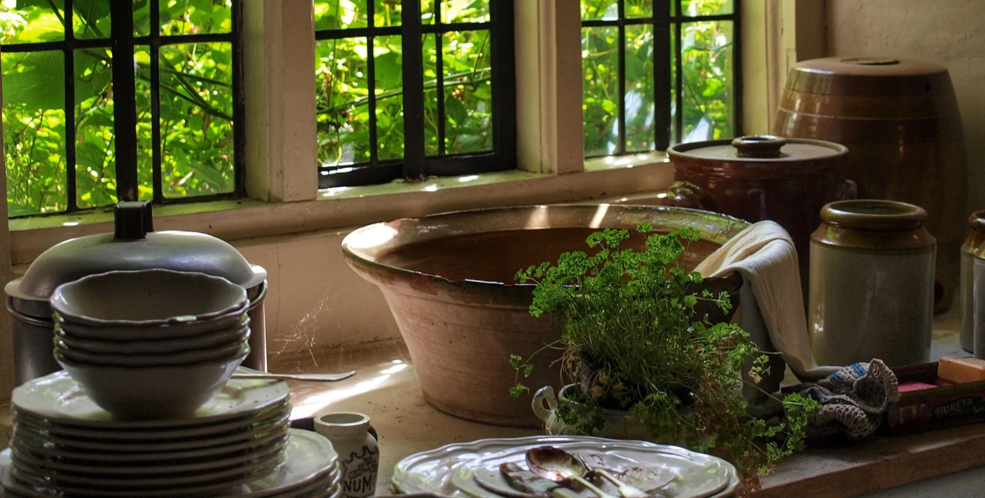 A rustic kitchen corner showcasing stacked crockery and herbs by a sunlit window, evoking warmth and charm.