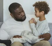 Content African American boy sitting on knees of caring black bearded father on couch in light living room at home