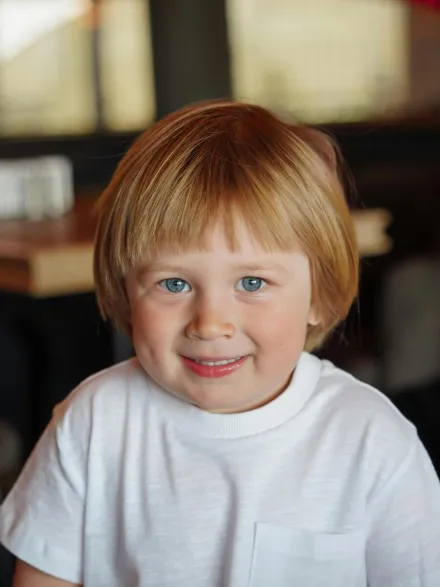 Close-up portrait of a cheerful blonde child smiling indoors, wearing a white t-shirt.