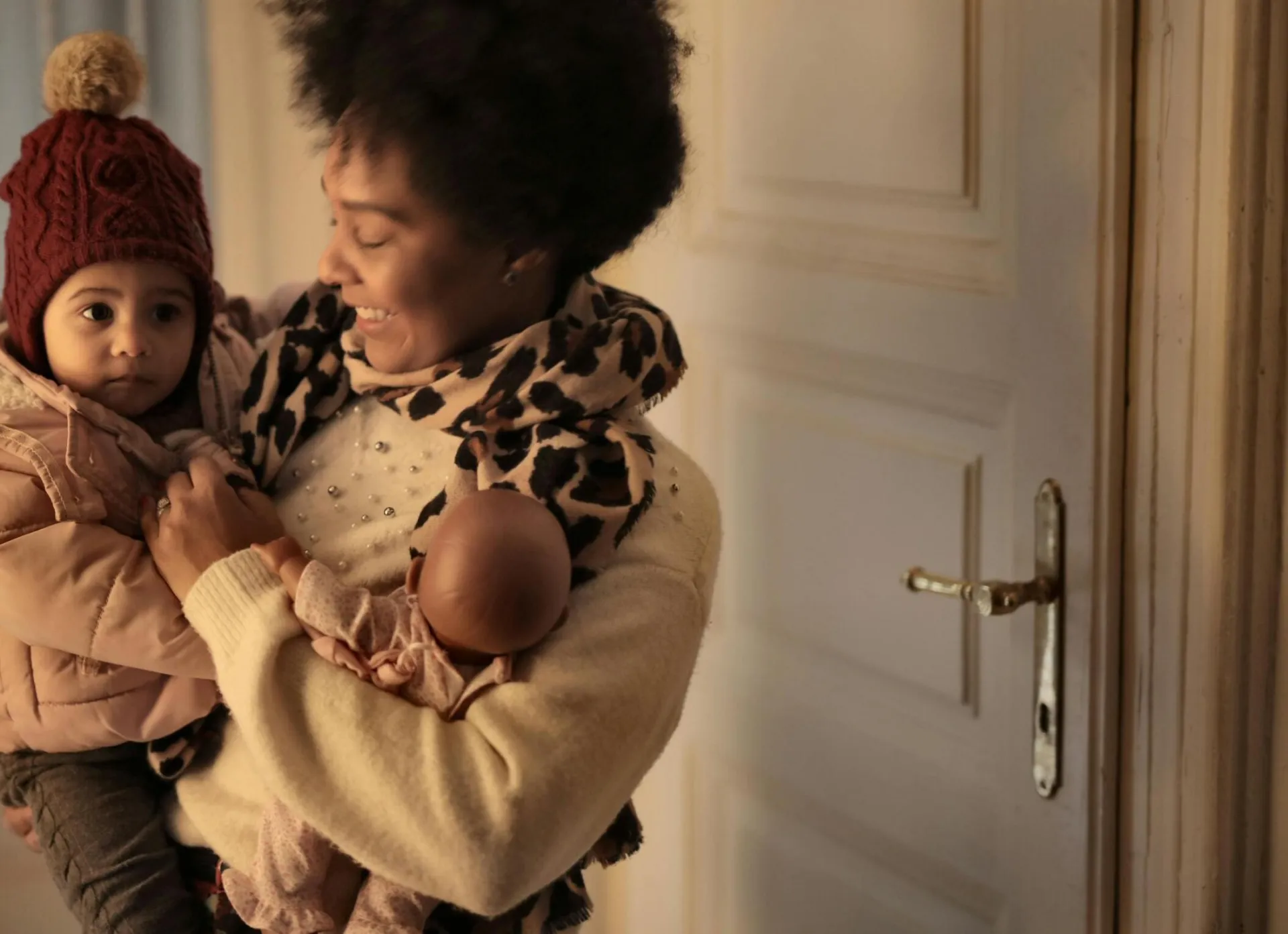 Cheerful ethnic woman hugging little kid while standing in room with toy and looking at clothes of baby while preparing to walk