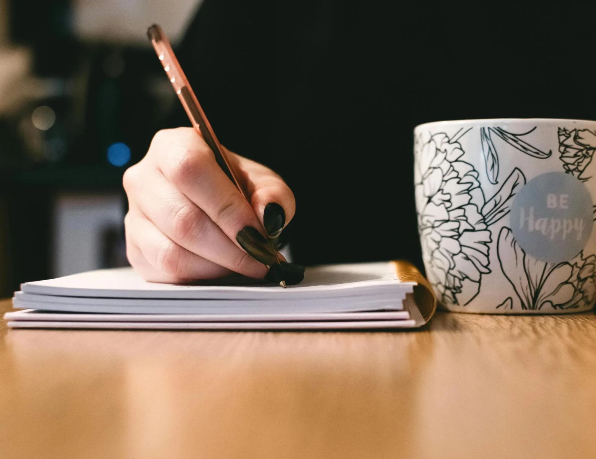 Person writing in a notebook with a floral ceramic mug on a wooden desk.