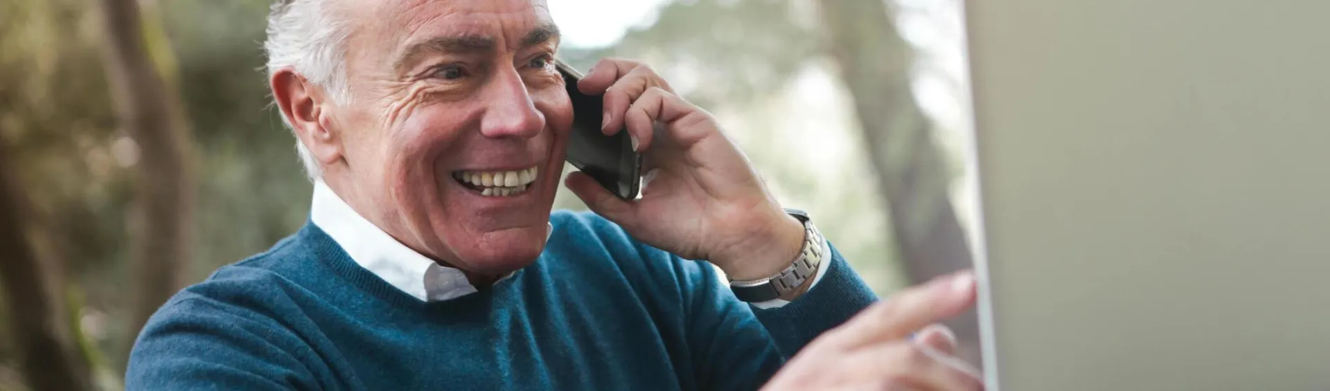 Cheerful senior man working remotely, talking on phone and using laptop outdoors.
