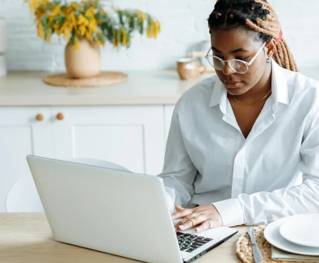 African woman in a kitchen setting working on a laptop at a dining table.
