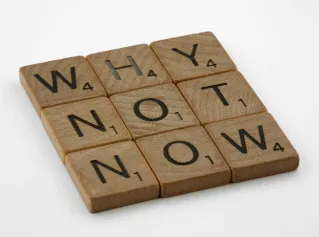 Wooden letter tiles form the motivating phrase 'Why Not Now' on a white background, encouraging action and decision-making.