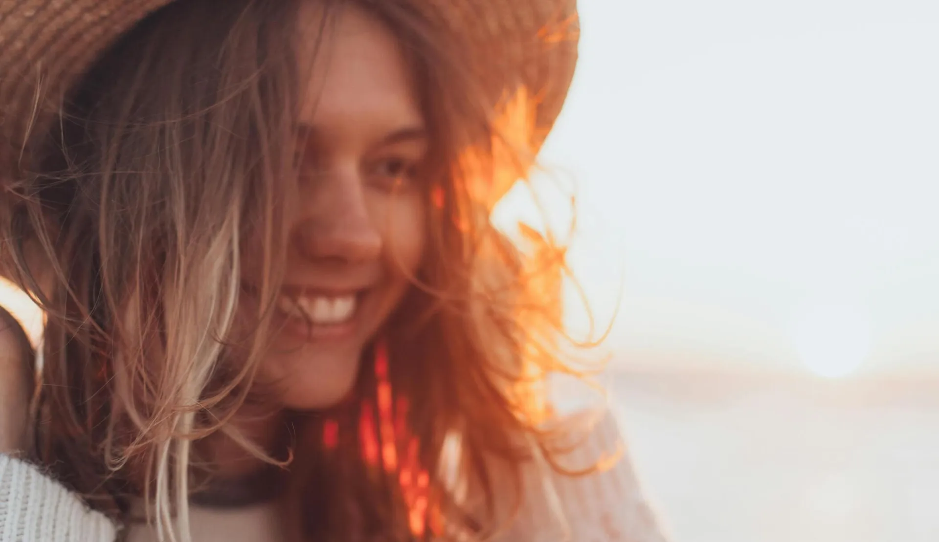 Joyful woman in a straw hat smiling by the seaside with sunset glow.