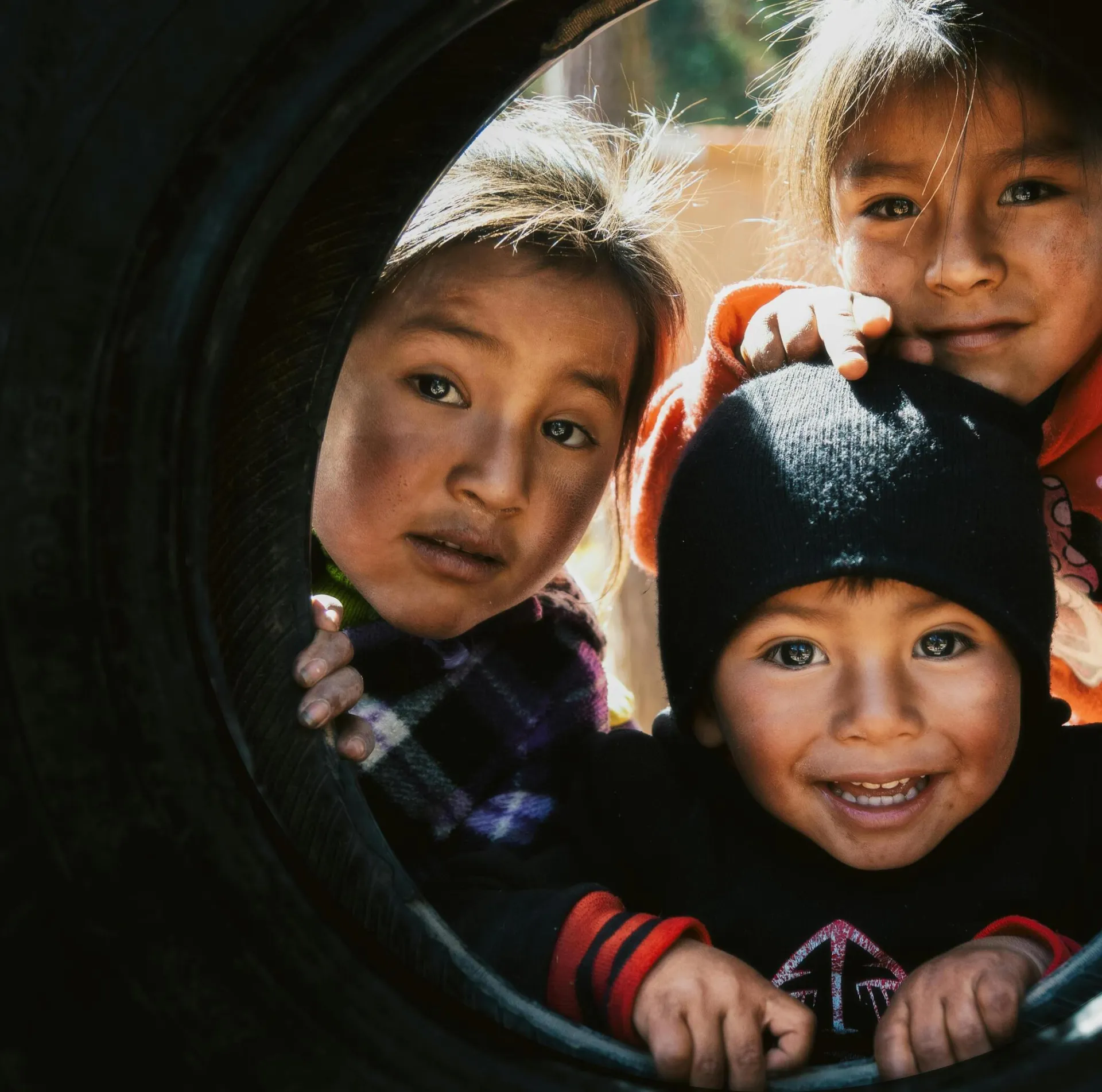 Three happy children play inside a tire swing outdoors in Cusco, Peru showcasing joy and friendship.