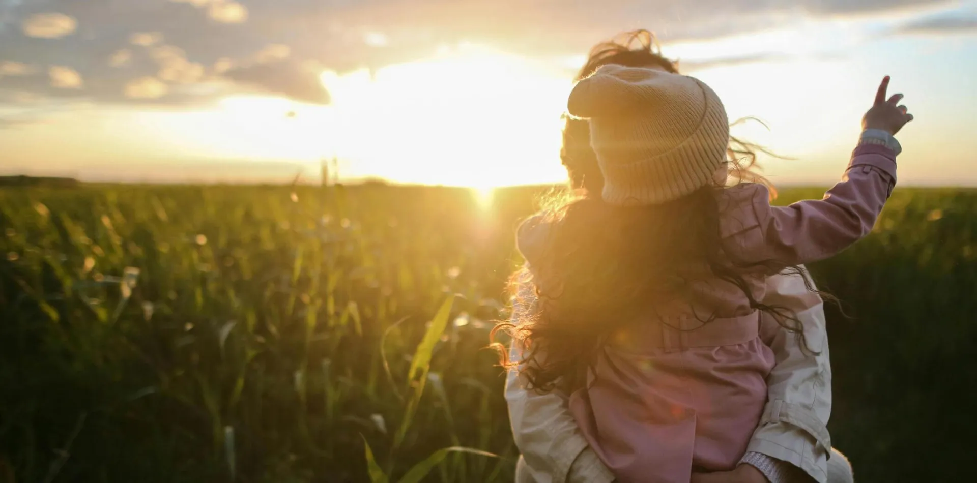 A mother and daughter embrace and point at the sunset in a grassy field.