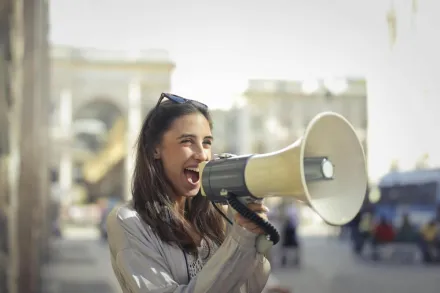 Cheerful young woman in a casual outfit shouting into a megaphone on a sunny day.