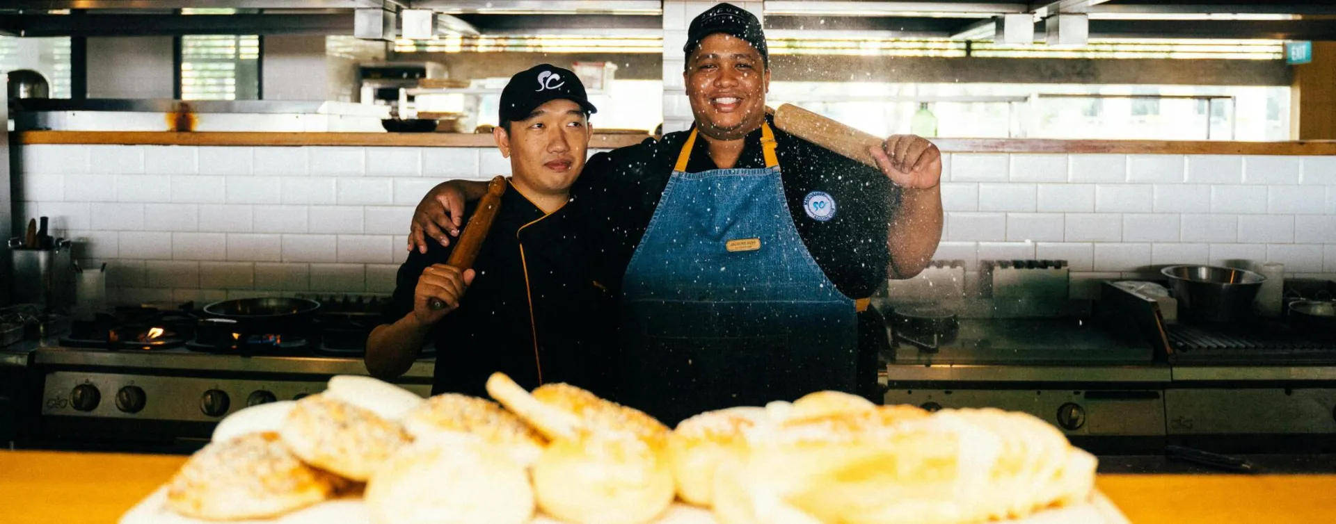 Two chefs in a restaurant kitchen with baked goods, smiling and holding a rolling pin.