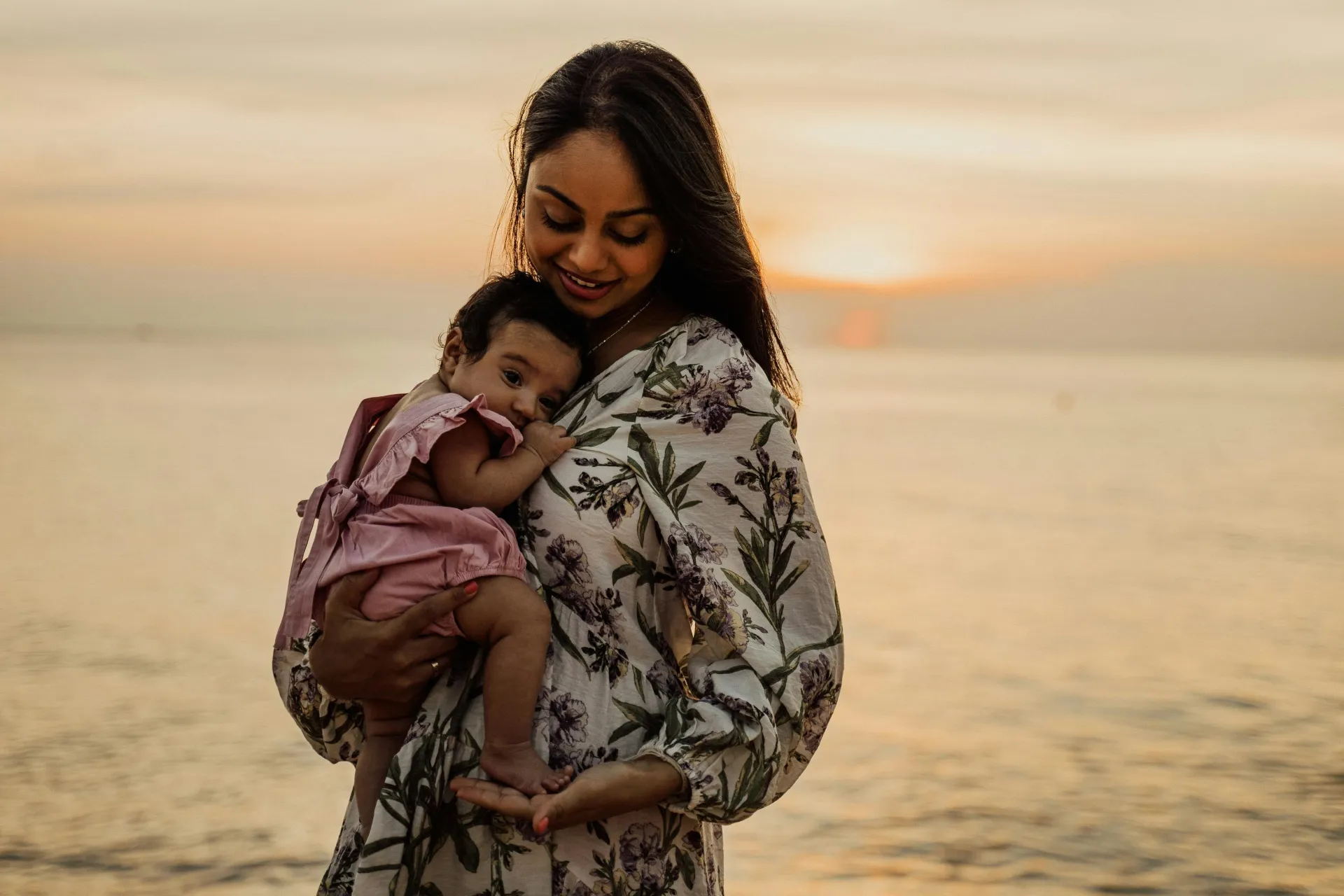 A serene moment of a mother holding her baby during sunset at the beach.