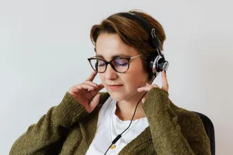 Content glad female wearing eyeglasses and headphones listening to good music and touching headset while sitting with eyes closed against white wall