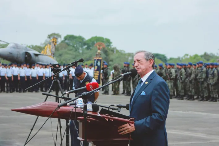 Side view of mature colonel with order standing near microphones on asphalt pavement behind army of soldiers near airplane monument under serene sky during national military holiday and looking away