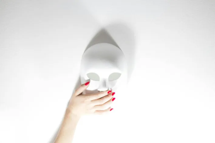 A hand with red nails holding a white face mask on a white background, creating a minimalistic vibe.