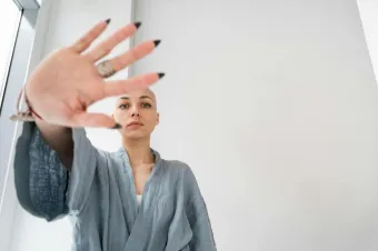 Bald woman in a robe making a stop gesture indoors, symbolizing resistance or caution.