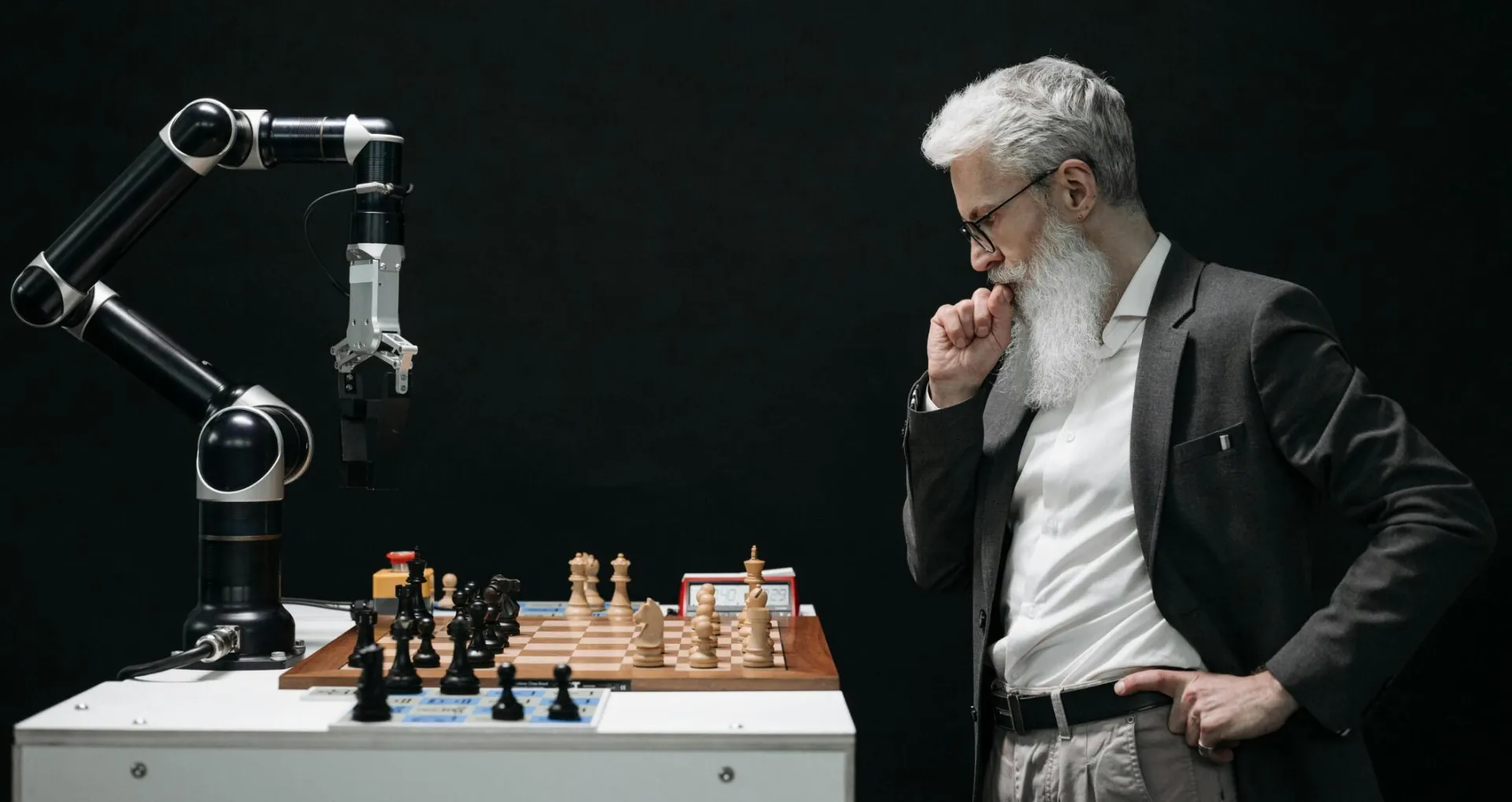 An elderly scientist contemplates a chess move against a robotic arm on a chessboard.
