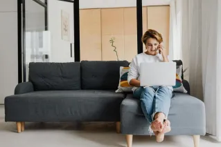 The Founder of The Pop In Concierge sitting barefoot on a sofa, working on a laptop and talking on the phone