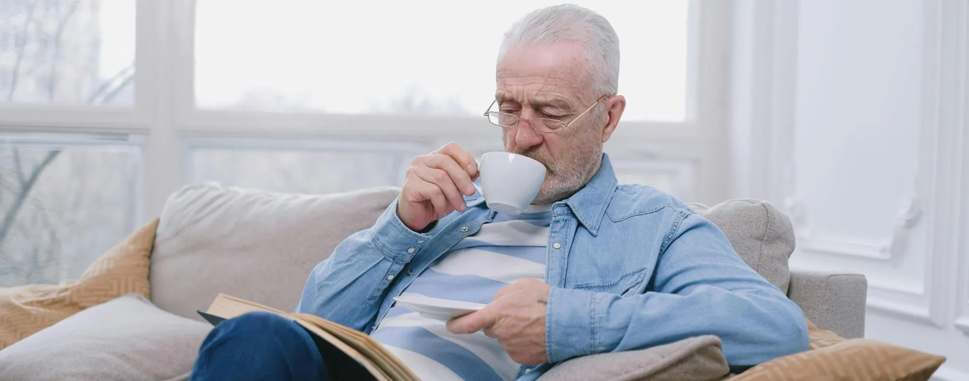 Elderly man enjoying a peaceful moment on sofa with a hot drink and book.