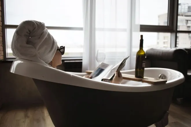 Woman relaxing in a bathtub with a towel wrapped head, reading and enjoying wine indoors.