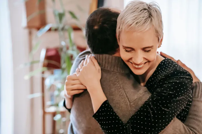 Positive gentle wife with toothy smile hugging husband while spending romantic time at home together