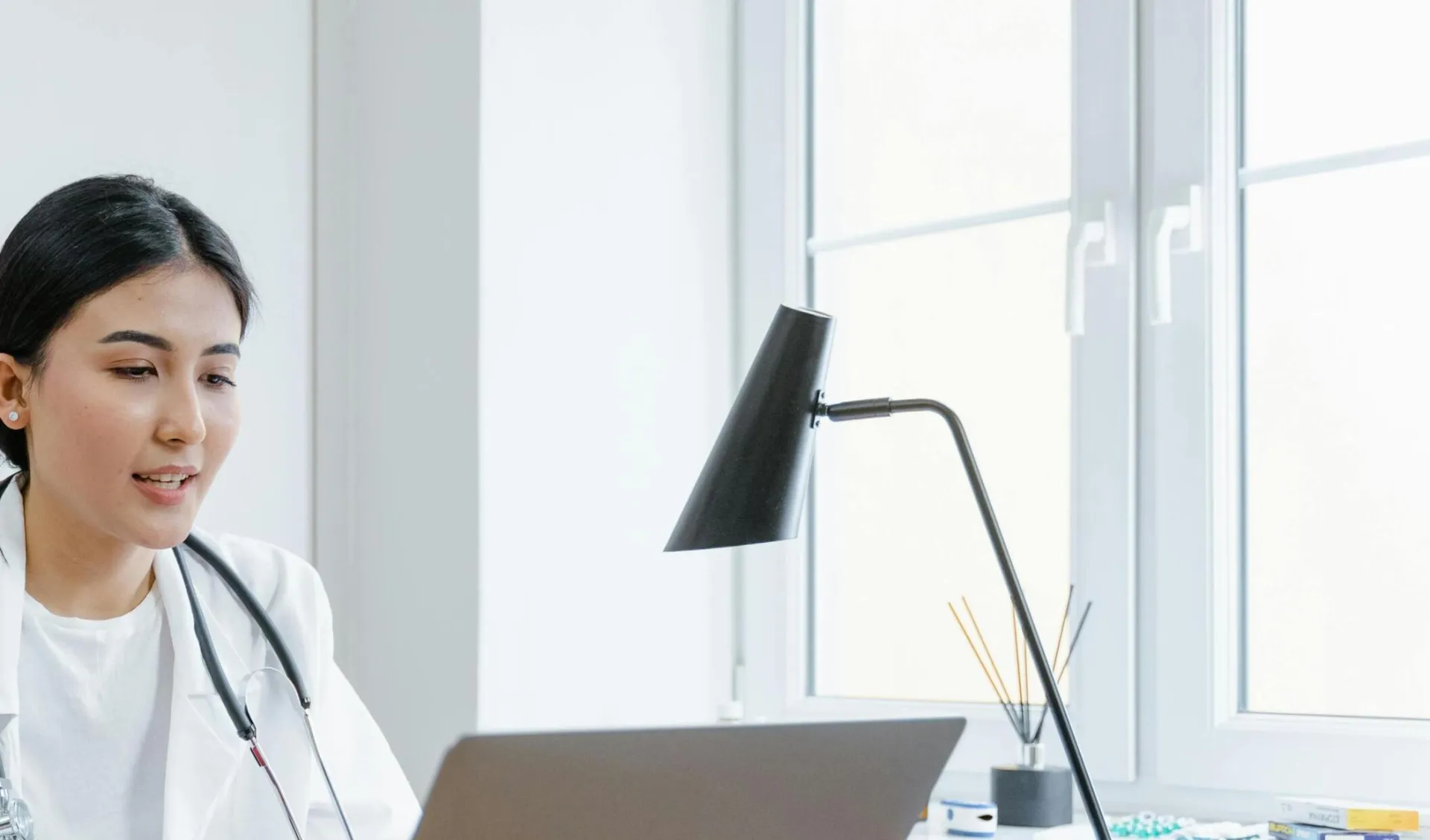A female doctor in a white coat uses a laptop for an online consultation from her office.