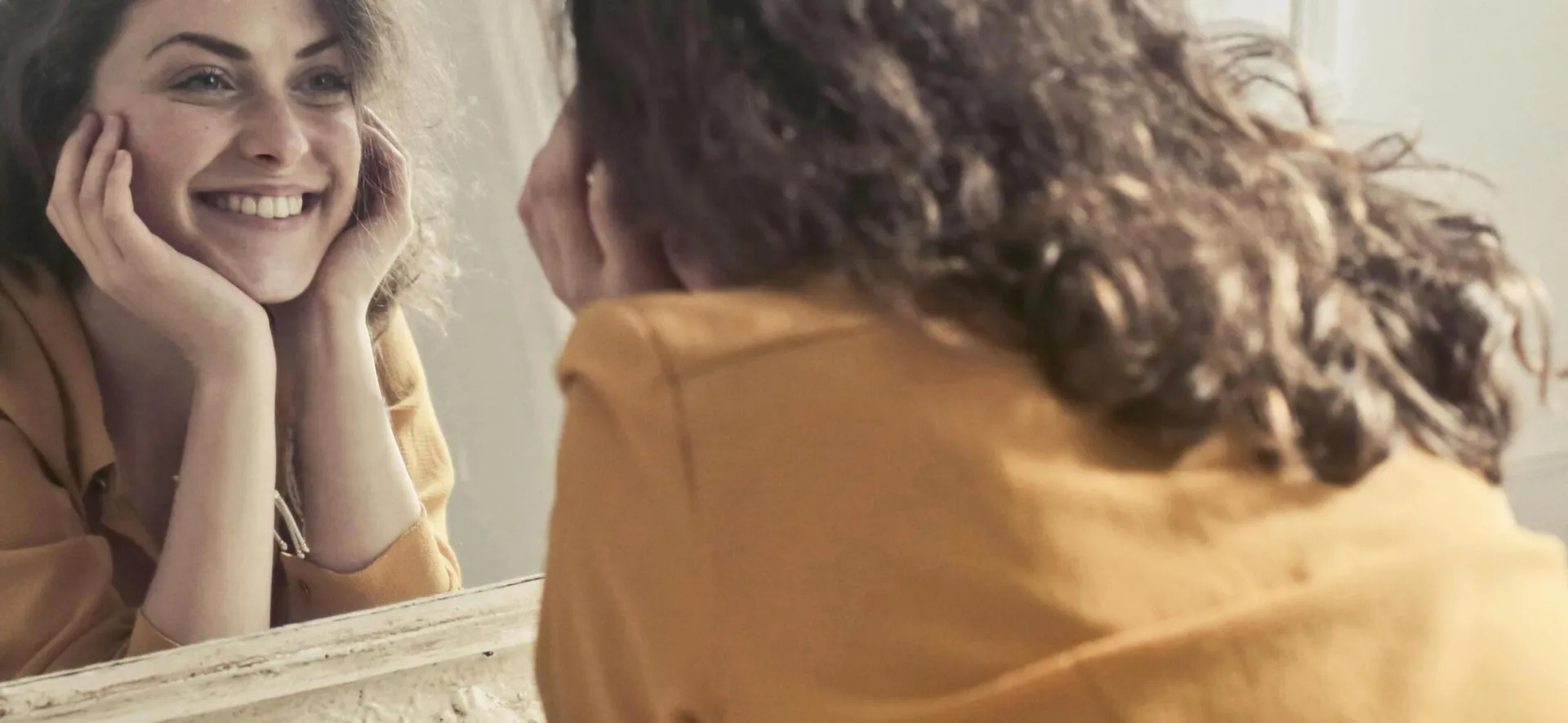 A cheerful woman smiles at her reflection in a vintage-style mirror, exuding positivity and warmth.