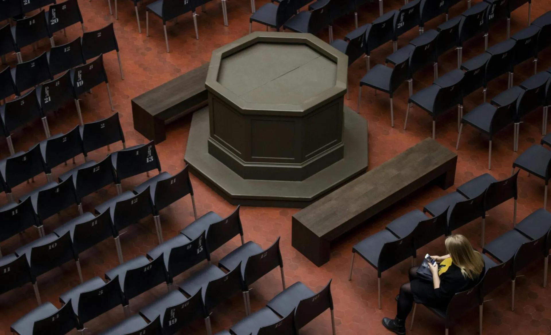 A solitary woman seated among rows of empty chairs in a spacious indoor auditorium.