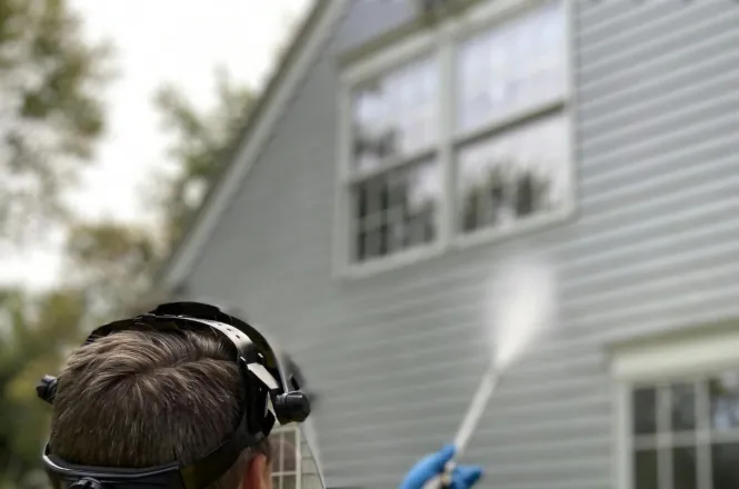 Person cleaning house siding with a pressure washer, wearing protective gear in a yellow raincoat.