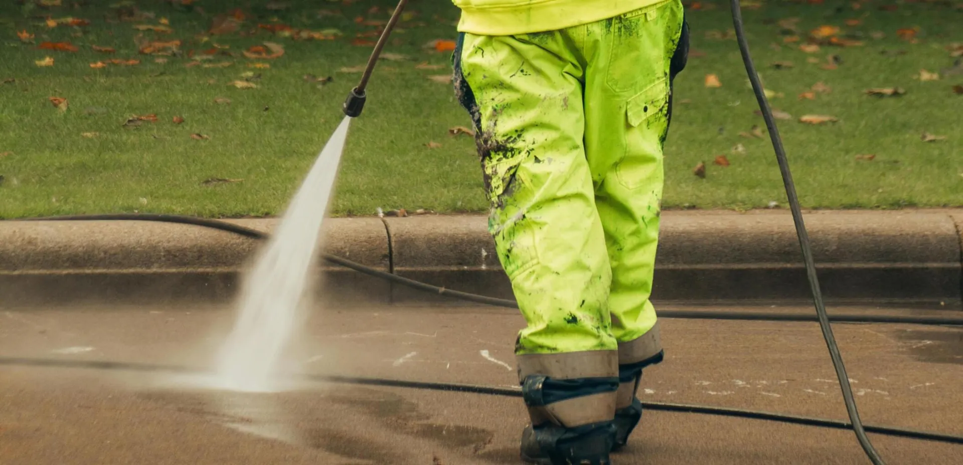 A city worker in high-visibility gear pressure washing a street corner.