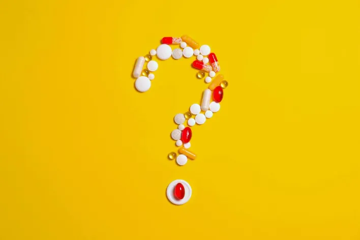 Assorted pills and capsules arranged in a question mark on a bright yellow background symbolizing medication inquiries.