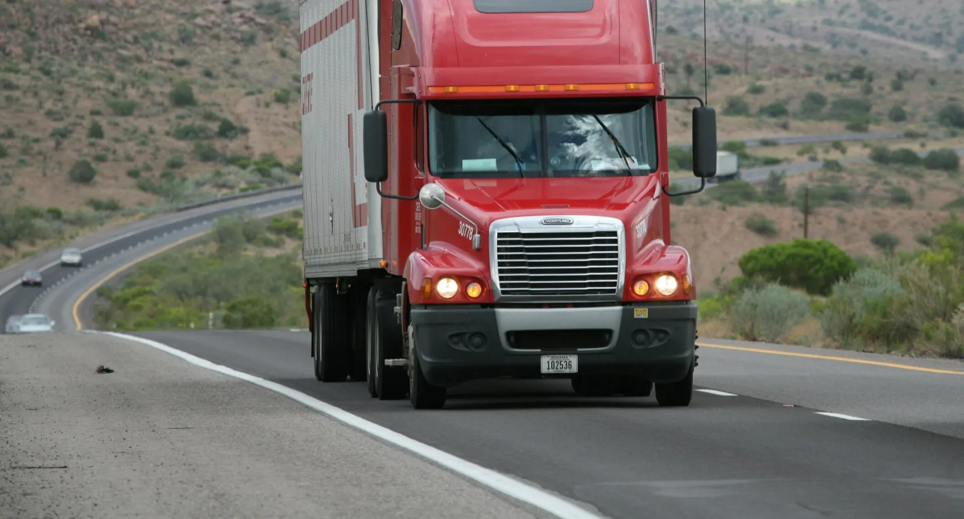 Red semi-truck driving through scenic highway in a mountainous landscape.