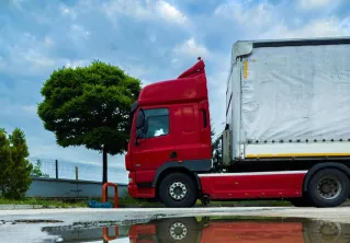 A parked red truck with a tarp-covered cargo is reflected in a puddle on an urban street, under a cloudy sky.