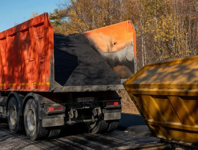 A dump truck unloading asphalt into a construction bin on a sunny autumn day.